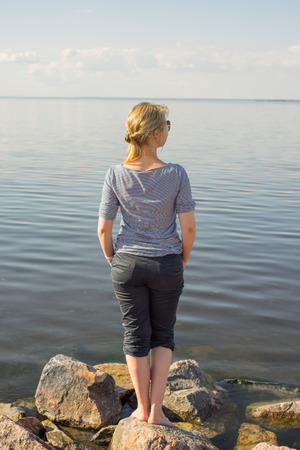 Beautiful young girl in a gray striped sweater with white hair stands on the stones against the background of the lakeの写真素材