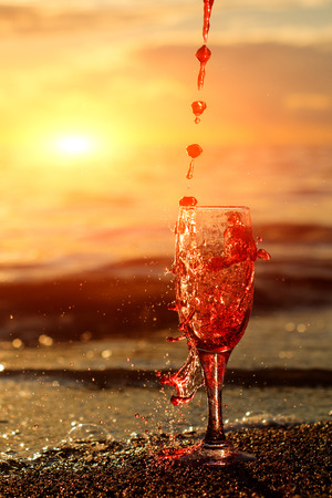 Romantic beach scene: glass of red wine at sunset near water line with splashesの写真素材