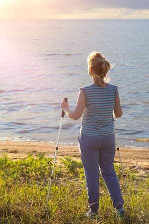 Beautiful young girl blonde in striped shirt without sleeves with hiking sticks looks at the sunset. Back viewの写真素材