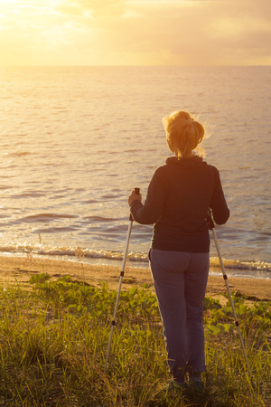 Beautiful young girl blonde in a dark sweater with hiking sticks looks at the sunset. Back viewの写真素材