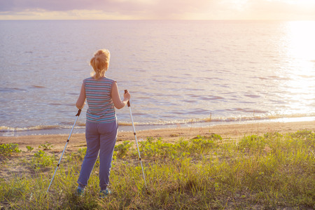 Beautiful young girl blonde in striped shirt without sleeves with hiking sticks looks at the sunset. Back viewの写真素材