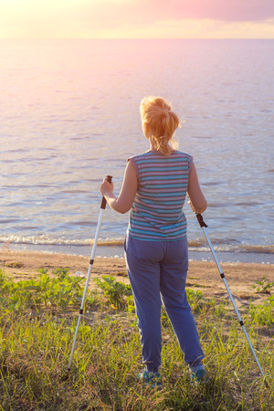 Beautiful young girl blonde in striped shirt without sleeves with hiking sticks looks at the sunset. Back viewの写真素材