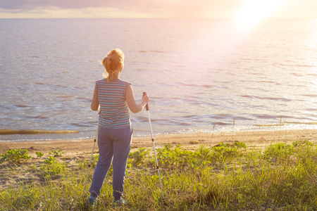 Beautiful young girl blonde in striped shirt without sleeves with hiking sticks looks at the sunset. Back viewの写真素材