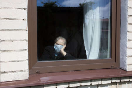An old grandmother in a protective mask looks out the window at self-isolation. Elderly woman at quarantine at home.の写真素材