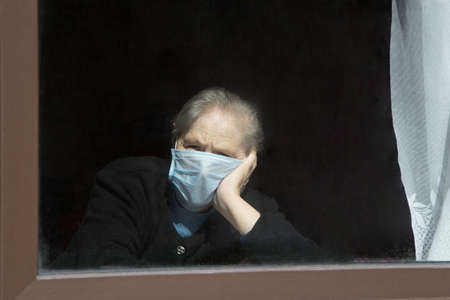 An old grandmother in a protective mask looks out the window at self-isolation. Elderly woman at quarantine at home.の写真素材