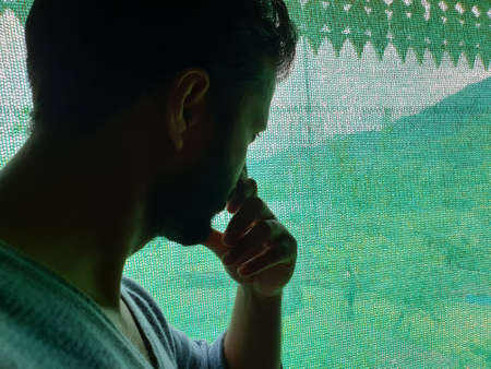A man is sad, helpless, depressed, in tension thinks on coronavirus.Corona or cover-19 stay home pandemic symptoms deadly virus fever cough covering mouth nose. Model looking outside through green netの写真素材