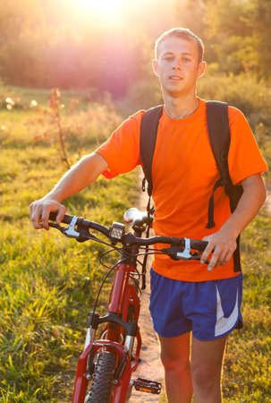 Happy man cyclist with bicycle walk a backpack behind shoulders illuminated by the rays of summer sunの写真素材