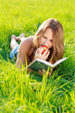 Happy beautiful woman with an apple in hand lying on the green grass and reading a book a sunny day against a background of green natureの写真素材