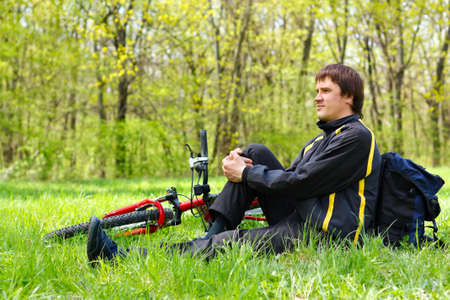 Happy man cyclist with bike and backpack sitting on green grass against the background of nature in spring on a sunny dayの写真素材