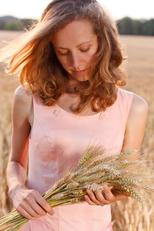 Young woman with ripe ears of wheat in the hands against the background of a wheat field in the eveningの写真素材