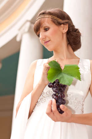 Portrait of beautiful greek woman with bunch of grapes in her hands against the background of a classical building with columnsの写真素材