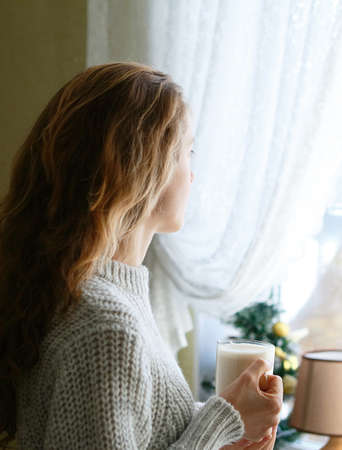 Young woman with a cup of tea in her hand looking through the windowの写真素材