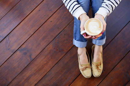Woman holding in hands cup of coffee with milk sitting on the wooden floor, top view pointの写真素材