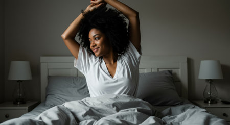 A relaxed African American woman with curly hair is stretching in bed with light grey sheets, smiling gently as she wakes up. Bedroom interior.の素材