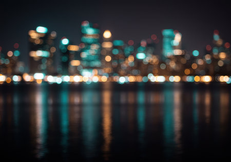 Defocused cityscape at night with blurred lights reflecting on water. Modern architecture with bokeh effect. Urban skyline with illuminated buildings.の素材