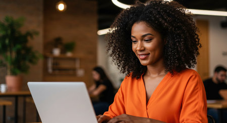 Happy African American woman with curly hair typing on a laptop in a modern coworking space. She is smiling while working on technology.の素材