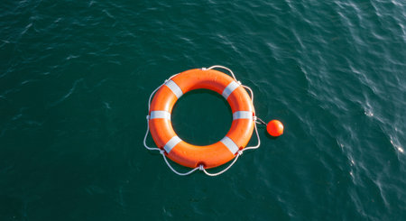 Orange lifebuoy with white stripes and rope floating on the calm, dark water surface. Symbol of safety, rescue, and help at sea.の素材