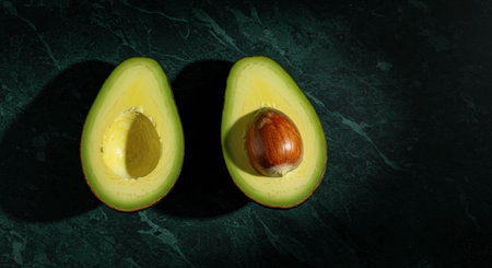 Two halves of a fresh, ripe avocado, one showing the large pit, arranged on a dark green marble surface. Concept of healthy eating and vegan food.の素材