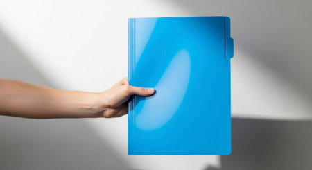 A fair skinned hand extends from the left, holding a vibrant blue plastic folder against a white background with natural shadows. Represents organization, data management, and professional documentation.の素材