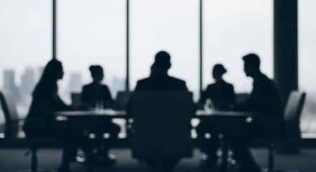 Blurred silhouettes of business professionals in a modern meeting room, gathered around a table with a city skyline visible through a large window. Represents teamwork and corporate discussion.の素材