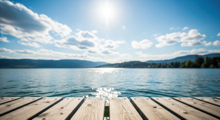 A sunlit wooden pier extends over calm lake water with sparkling reflections, under a bright blue sky with white clouds and distant mountains.の素材