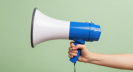 A human hand holds a blue and white megaphone, symbolizing communication, public address, and making an announcement. The device is ready to amplify a message.の素材