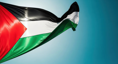 A vibrant palestine flag with black, white, green stripes and a red triangle waving against a clear blue sky. The fabric shows movement and texture.の素材
