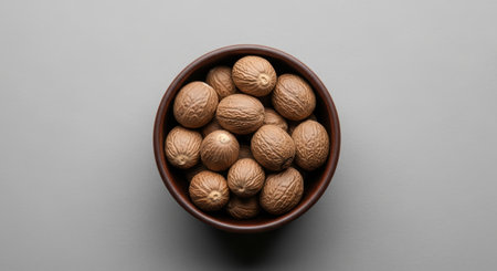 A top down studio shot of a dark brown ceramic bowl filled with whole nutmegs on a plain grey background. This spice is commonly used in cooking and baking for its aromatic flavor, representing ingredients and natural food.の素材