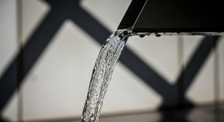 A close up view of a continuous stream of crystal clear water flowing from a modern, sleek black faucet. Water droplets are visible on the faucet edge, highlighting the liquid movement and purity.の素材
