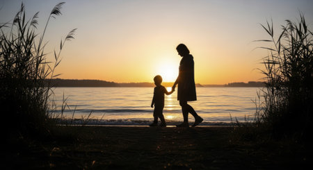 Silhouettes of a mother and her son holding hands, strolling along the water edge at sunset. golden light reflects on the tranquil lake, framed by tall reeds.の素材