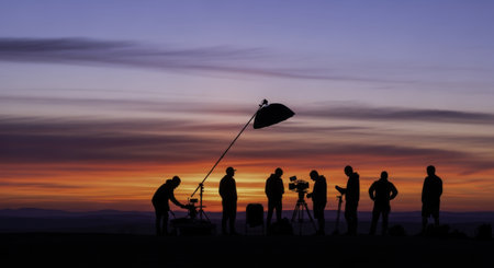Professional film crew silhouettes working outdoors with camera and lighting equipment against a vibrant sunset sky. capturing a cinematic production scene.の素材