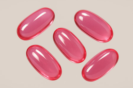 Five glossy translucent pink gel capsules, oval shaped, for health, medicine, and beauty supplements, on a neutral background.の素材