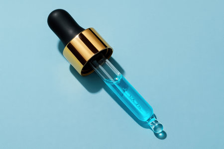 Clear glass dropper with vibrant blue liquid, black rubber bulb, and gold metal collar on a light blue surface, ideal for cosmetic and scientific concepts.の素材