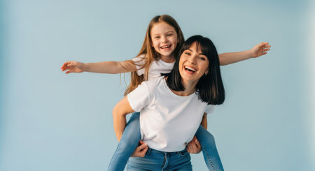Joyful mother giving a piggyback ride to her happy daughter, both smiling and laughing in white t shirts and blue jeans, celebrating family bond.の素材