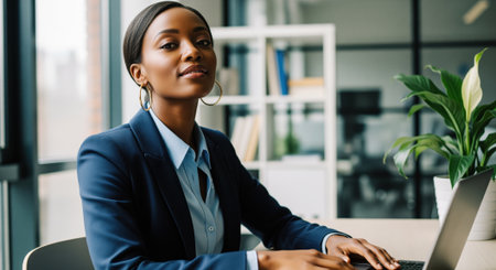 Stylish african businesswoman confidently poses at her desk in a modern office, working on a laptop. professional setting with natural light, reflecting success and corporate environment.の素材