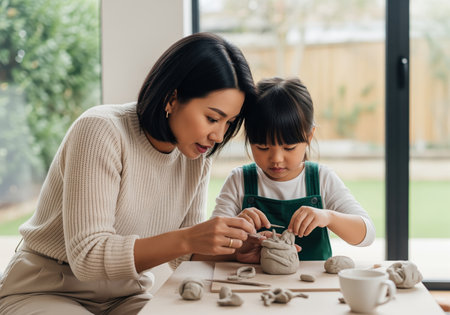 Asian mother and young daughter focused on sculpting clay figures with tools at a table. creative family activity, bonding, and artistic development.の素材
