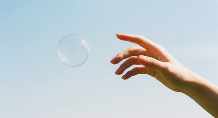 Female hand with manicured nails reaching for a floating, shimmering soap bubble in a bright blue sky. symbolizes lightness, freedom, and aspiration.の素材