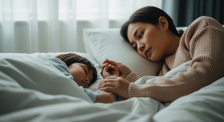 Asian mother gently holds her sleeping child hand in a cozy bed. depicts maternal bond, care, comfort, and peaceful rest.の素材