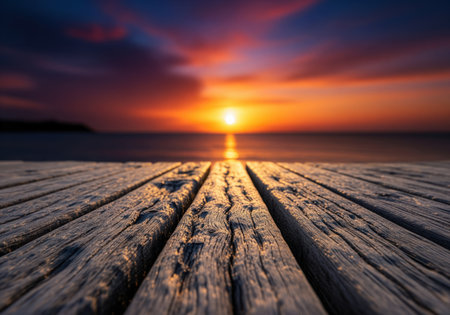 Weathered wooden planks of a pier extend towards a vibrant sunset over the tranquil ocean. the sky displays warm orange, red, and purple hues, with the sun reflecting on the calm water surface.の素材