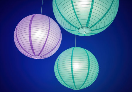 Three illuminated spherical paper lanterns, one purple and two teal, hang at different heights against a vibrant deep blue background. modern decorative lighting for events or home decor.の素材