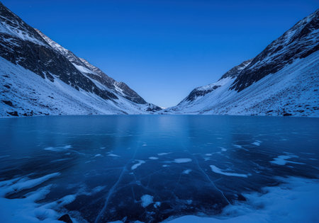 Frozen alpine lake with a glass like surface reflecting the deep blue twilight sky, nestled between steep, snow covered mountain slopes. a serene and cold winter landscape.の素材
