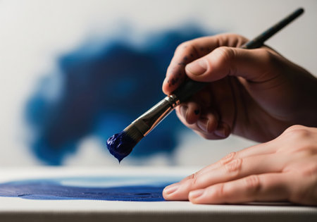 Artist hand holding a paintbrush loaded with rich blue paint, actively creating an artwork on a white canvas. focus on the creative process and artistic expression.の素材