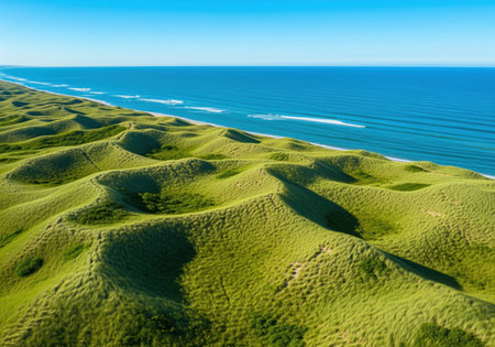 Expansive aerial landscape featuring rolling green dunes covered in grass adjacent to a pristine beach and deep blue sea under a clear sky. ideal for travel, nature, and environmental themes.の素材
