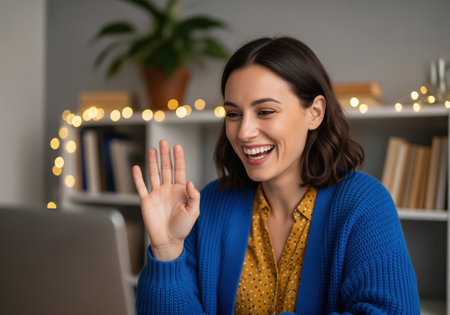 Cheerful young woman participates in a virtual meeting or social call, smiling widely and waving at her laptop screen from her home office setup.の素材
