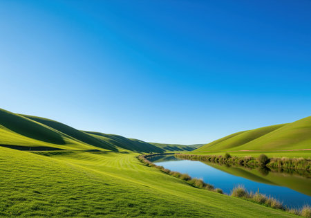 Lush green rolling hills and manicured slopes surround a winding river reflecting the clear blue sky, creating a pristine, idyllic natural landscape.の素材