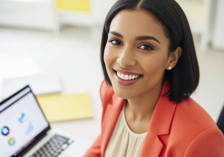 Confident young businesswoman with dark hair wearing an orange blazer smiling brightly at the camera while working on a laptop in a bright, modern office environment. represents success, professionalism, and corporate leadership.の素材