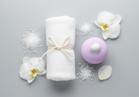 Rolled white cotton towel tied with twine, purple bath bomb, white bath salt crystals, and delicate orchid flowers arranged for a relaxing spa treatment on a gray surface.の素材