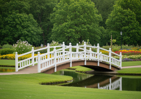 White wooden footbridge arching over a calm pond in a meticulously maintained public park or botanical garden, surrounded by vibrant green grass, lush trees, and colorful flower beds.の素材