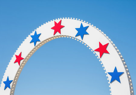 Section of a white amusement park arch decorated with alternating red and blue stars and illuminated by small bulbs, set against a clear, sunny blue sky.の素材