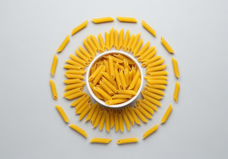 Dry penne pasta pieces meticulously arranged in a circular, decorative pattern surrounding a white bowl filled with more pasta. flat lay, top view on a light gray surface. represents italian cuisine and cooking ingredients.の素材